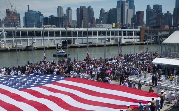 A large ceremonial American flag is being displayed by military personnel in white Navy uniforms and khaki Marine uniforms at what appears to be a waterfront pier ceremony in New York City. A large crowd of attendees, including civilians and military members, is gathered behind the flag. The urban skyline and a body of water are visible in the background, suggesting a location along the Hudson River.