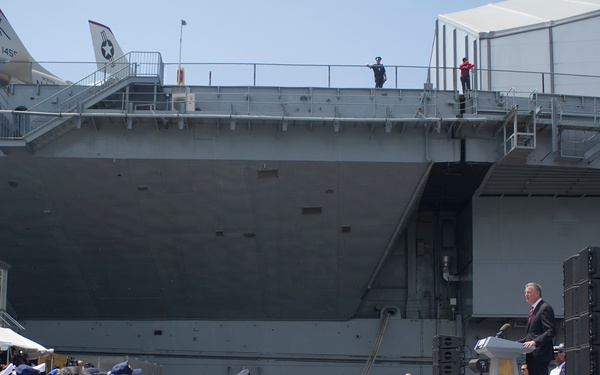 An outdoor patriotic ceremony is taking place alongside a large naval vessel, likely an aircraft carrier, with a speaker at a podium addressing a seated audience. The event features red, white, and blue bunting decorations along the stage area, uniformed naval personnel in formation, and security personnel standing nearby. Aircraft are visible on the flight deck of the carrier above.