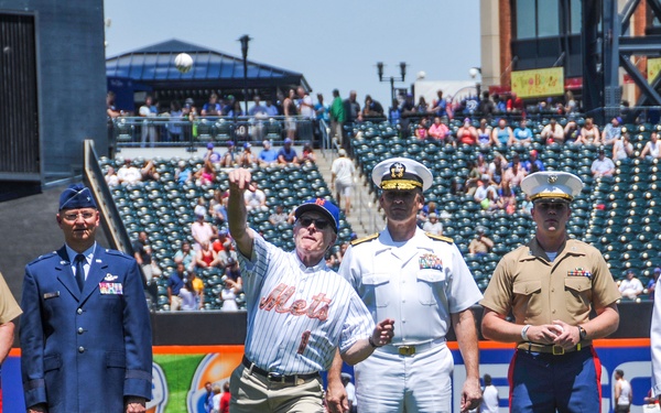A ceremonial first pitch is being thrown at a New York Mets baseball game, with the pitcher wearing a Mets jersey and cap. He is flanked by five military personnel representing multiple branches of the U.S. Armed Forces, suggesting a military appreciation or Armed Forces Day event. The game is being played at Citi Field with a large crowd visible in the background.