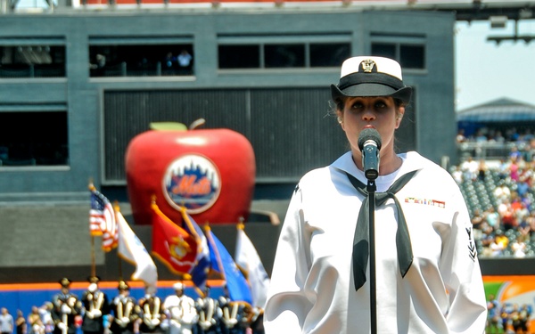 A U.S. Navy sailor in white dress uniform stands at a microphone on the baseball field at Citi Field, home of the New York Mets, appearing to sing the national anthem. A color guard with multiple military branch flags is visible in the background near the pitcher's mound. The iconic large red apple with the Mets logo is prominently displayed in the outfield.