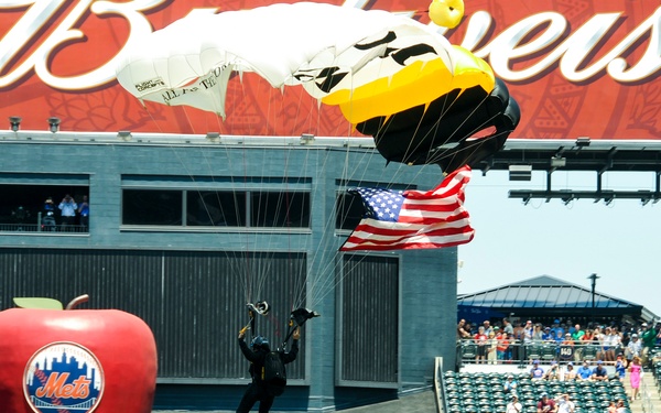 A parachutist carrying an American flag is descending into Citi Field, home of the New York Mets, during what appears to be a pre-game ceremonial skydive. The jumper is wearing a black jumpsuit and is accompanied by a black-and-yellow parachute, landing near the iconic large red apple sculpture in the outfield. The stadium is partially filled with spectators watching the event.