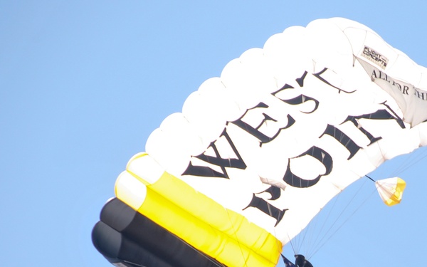 A parachutist is descending under a large white, black, and yellow canopy bearing the text 'WEST POINT'. The jumper is carrying an American flag while in flight against a clear blue sky. This appears to be a ceremonial or demonstration skydive, likely associated with the United States Military Academy at West Point.