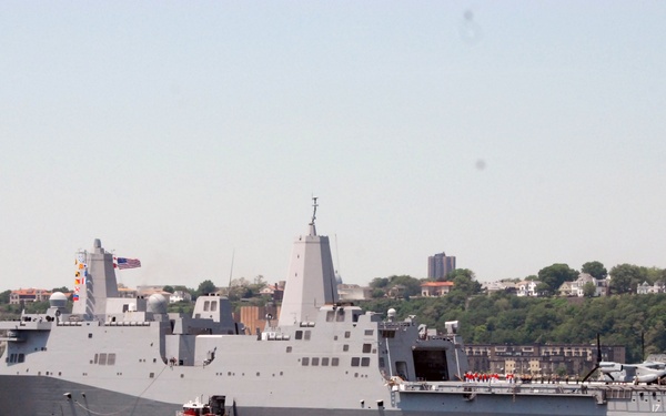 A large U.S. Navy amphibious transport dock ship, bearing the hull number 17, is underway on a waterway with an urban shoreline visible in the background. A red tugboat is positioned alongside the warship, likely providing assistance, while a smaller blue-and-white patrol boat operates nearby. An American flag is visible flying from the ship's mast.