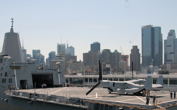 A U.S. Navy amphibious transport dock ship bearing hull number 17 and the name 'San Antonio' is docked at a pier with a dense urban skyline visible in the background, consistent with New York City. The ship's flight deck is visible with at least one tiltrotor aircraft, identified as an MV-22 Osprey, parked on it. Several personnel are visible on the deck and pier area.
