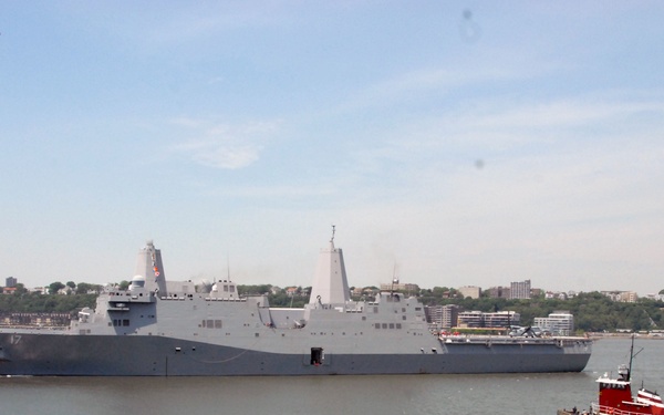 A large U.S. Navy amphibious transport dock ship, bearing the hull number 17, is underway on a wide river or harbor with an urban shoreline visible in the background. Two tugboats — one red and yellow on the starboard side and one smaller vessel on the port side — are assisting or escorting the warship. The setting appears to be a major metropolitan waterway, likely New York Harbor based on the skyline and geography.