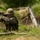 A soldier in camouflage uniform and helmet is positioned in a field fortification or trench, appearing to cover their ears or communicate via radio. A mortar tube on a bipod mount is set up to the soldier's right in a prepared firing position. The setting appears to be a military training exercise or field operation in a wooded, grassy area.