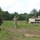 A military training exercise appears to be underway in an open field with mixed forest in the background. Two soldiers on foot are directing or coordinating movement, while an M113 armored personnel carrier (APC) with crew members visible in the hatch operates nearby. The soldier on the left wears a Polish military uniform with a visible Polish flag patch, while the center soldier wears US-pattern MultiCam/OCP camouflage, suggesting a joint multinational exercise.