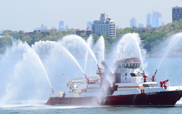 A red and white fireboat, visibly labeled 'FIREFIGHTER II' and 'FIRE', is navigating open water while simultaneously deploying multiple high-pressure water cannons in dramatic arcing sprays. The vessel appears to be on the New York Harbor or a similar urban waterway, with a densely treed shoreline, residential and commercial buildings, and a distant city skyline visible in the background. This appears to be a demonstration or ceremonial display rather than an active firefighting operation.