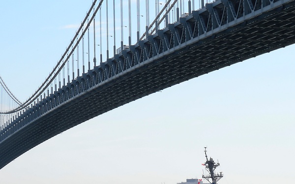 A U.S. Navy Arleigh Burke-class guided-missile destroyer is passing under a large suspension bridge, consistent with the Verrazzano-Narrows Bridge in New York. The warship appears to be participating in a fleet review or transit event, with personnel visible lining the deck rails. A smaller vessel and what appears to be a submarine or second naval vessel are also visible in the waterway.