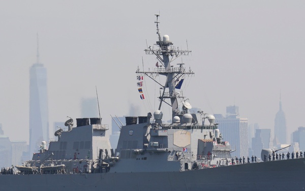 A U.S. Navy Arleigh Burke-class guided-missile destroyer, bearing hull number 55, is underway on open water with a large city skyline visible in the background. The skyline is identifiable as New York City, with One World Trade Center prominently visible. Crew members are visible manning the rails along the deck, suggesting a formal arrival or departure ceremony.
