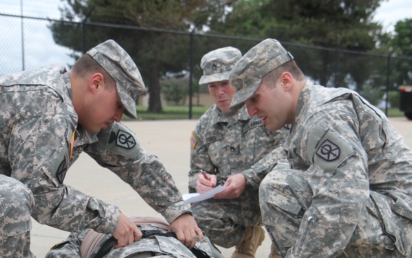 Three U.S. Army soldiers in ACU (Army Combat Uniform) are conducting a medical training exercise outdoors on a paved surface. Two soldiers are attending to a fourth soldier lying on a litter/stretcher, while a third soldier observes and takes notes on a clipboard. The setting appears to be a military installation with vehicles and a chain-link fence visible in the background.