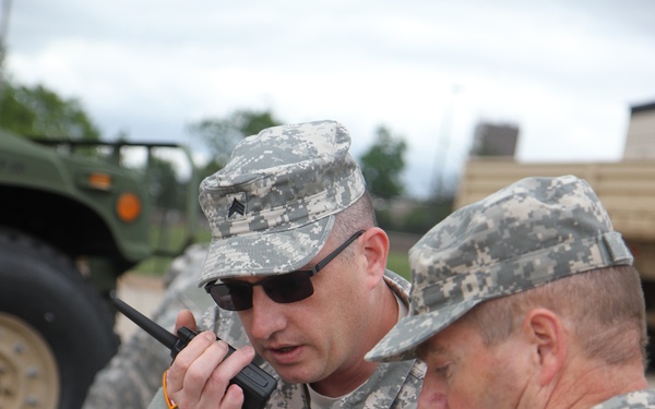 Two U.S. Army soldiers in ACU (Army Combat Uniform) digital camouflage are engaged in coordinated communication activities in an outdoor military setting. The soldier on the left is speaking into a handheld radio while the soldier on the right takes notes on a small notepad. Military vehicles are visible in the background.