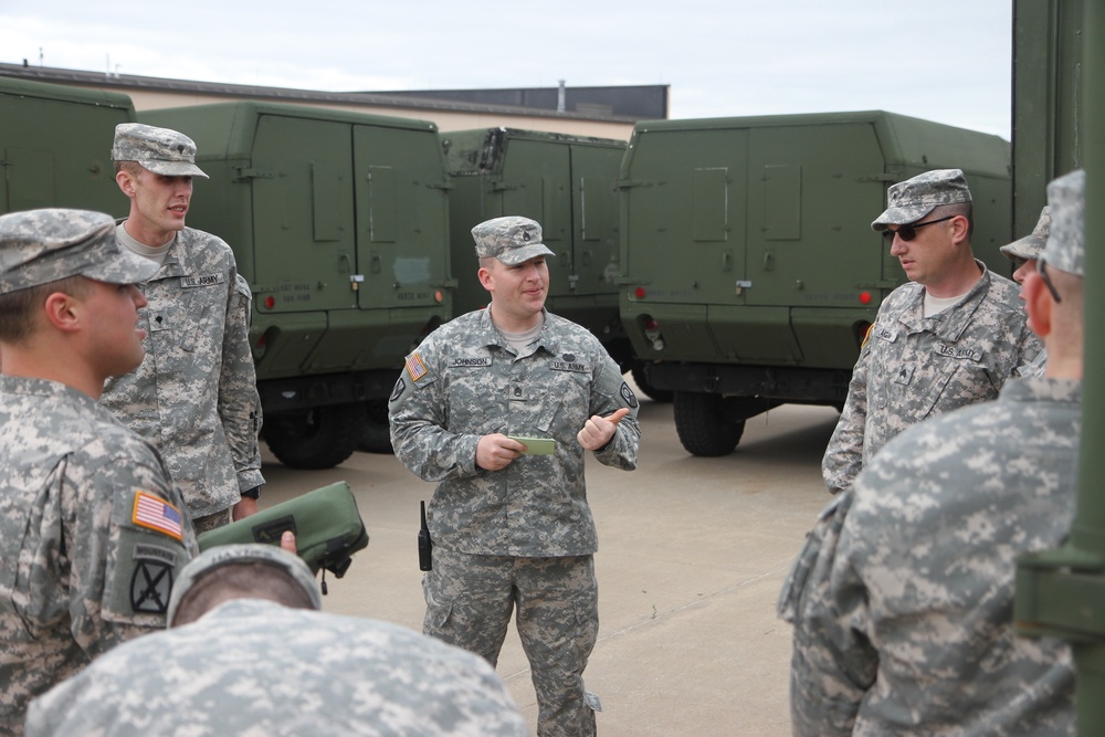 A group of U.S. Army soldiers in ACU (Army Combat Uniform) digital camouflage are gathered in a motor pool or staging area. A soldier identified by nametape as 'JOHNSON' appears to be conducting a briefing or instruction session while holding a small notepad. Several olive drab military vehicles are parked in the background.