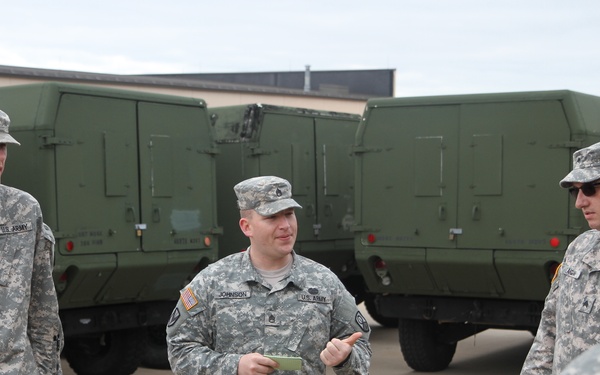 A group of U.S. Army soldiers in ACU (Army Combat Uniform) digital camouflage are gathered in a motor pool or staging area. A soldier identified by nametape as 'JOHNSON' appears to be conducting a briefing or instruction session while holding a small notepad. Several olive drab military vehicles are parked in the background.