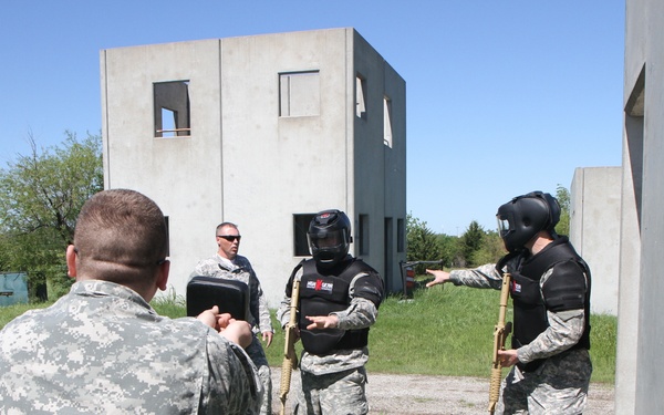 Military personnel are conducting a close-quarters combat or combatives training exercise at what appears to be a mock urban training facility. Two soldiers wearing protective gear including helmets, body armor, and holding padded training weapons are facing an instructor in ACU (Army Combat Uniform) camouflage. Additional personnel are observing from the side, one holding a clipboard or tablet.