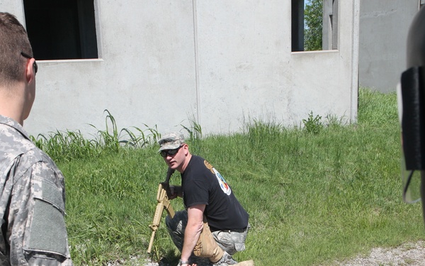 The image shows what appears to be a military training exercise involving tactical casualty care or combat lifesaver training. An instructor in a black shirt and patrol cap is kneeling over a person lying on the ground in ACU (Army Combat Uniform) pattern, demonstrating a medical or tactical procedure. Two other individuals in military uniforms are observing the demonstration near a concrete training structure.