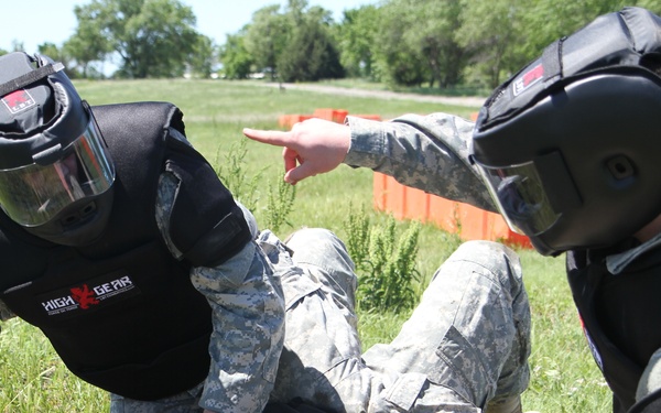 Two individuals in military camouflage uniforms and High Gear protective training equipment are conducting a ground control/detainment drill on a third person lying on grass. The two standing/kneeling personnel wear full protective helmets with face shields and padded body armor, indicating a combatives or law enforcement training exercise. One trainer is pointing, suggesting instruction is occurring simultaneously with the physical drill.