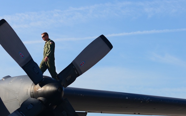 A military personnel member in a green flight suit is standing on the wing of a large military turboprop aircraft, near one of its large multi-blade propeller assemblies. The image is taken from a low angle, emphasizing the scale of the aircraft's wing and propeller against a clear blue sky. The scene appears to depict a pre-flight or post-flight inspection walkthrough.