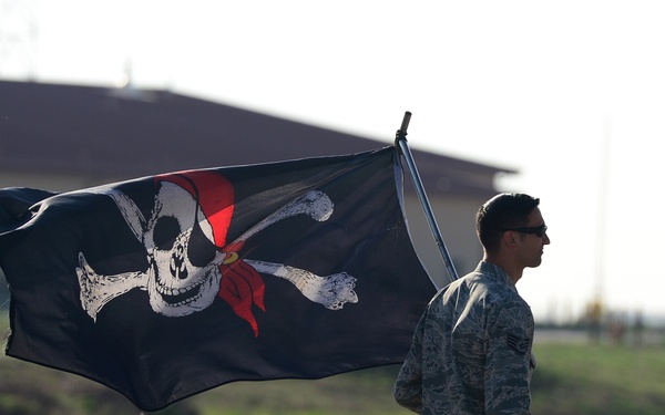 A military service member in camouflage uniform carries a large Jolly Roger (pirate) flag across an open field near a military installation. The flag features the classic skull-and-crossbones design with a red bandana accent. The setting appears to be a military base or airfield, likely during a ceremonial or informal event.