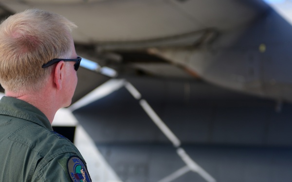 A military aviator in an olive green flight suit is conducting a pre-flight or post-flight walk-around inspection of a large military aircraft. The individual is viewed from behind, facing the underside and landing gear area of the aircraft. The setting appears to be a military flight line or tarmac.