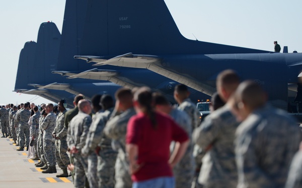 A large formation of military personnel, predominantly in US Air Force digital camouflage uniforms (ABUs), are lined up on an airfield tarmac alongside multiple large military aircraft. One civilian individual in a red shirt and blue pants stands among the uniformed personnel. The scene appears to be a formal ceremony or dignified transfer event on a military flight line.