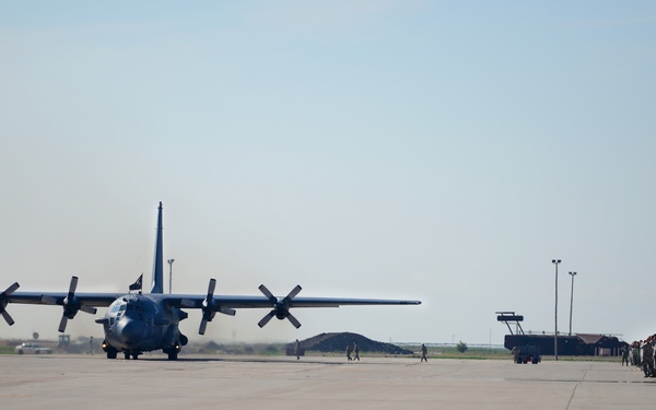 A military turboprop transport aircraft, consistent with a C-130 Hercules, is taxiing on a flight line apron at a military air base. A formation of uniformed military personnel is lined up on the right side of the tarmac, suggesting a formal arrival or departure ceremony. Additional aircraft tails are partially visible on the far right, indicating this is an active military flight line.