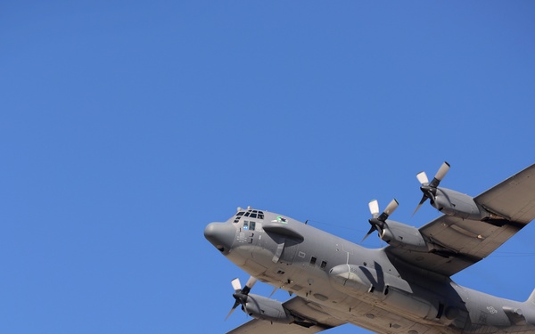 A military turboprop transport aircraft, consistent with a Lockheed C-130 Hercules variant, is photographed in flight against a clear blue sky. The aircraft is painted in a gray military finish and appears to be in a slight bank, offering a clear view of its underside and wing configuration. The image is sharp and well-exposed, capturing significant detail of the airframe.