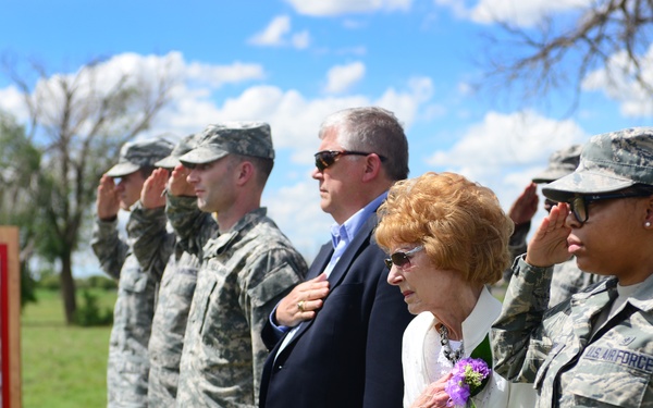 A formal outdoor military ceremony is taking place, with uniformed Air Force and Army personnel standing at salute alongside two civilians. An elderly woman in white with a floral corsage and a man in a suit stand among the military members in a show of respect. A display board with photographs is visible to the left, suggesting a dedication or commemorative event.