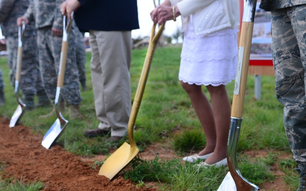 A military groundbreaking ceremony is taking place on a grassy field with red clay soil. A line of participants, including military personnel in camouflage uniforms and civilians, are holding ceremonial shovels along a freshly turned row of earth. One distinctive gold-painted shovel stands out among the otherwise silver ceremonial shovels.