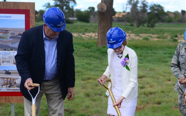 A groundbreaking ceremony is taking place at Cannon AFB for a clinic replacement facility. Four participants — two in civilian attire and two in U.S. Air Force camouflage uniforms — are using ceremonial shovels to break ground. A display board in the background shows architectural renderings labeled 'Cannon AFB Clinic Replacement.'