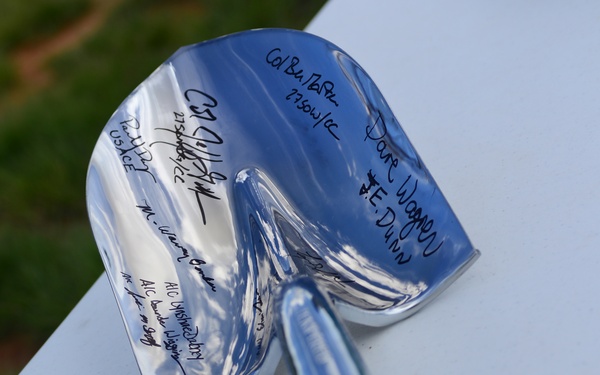 A polished ceremonial chrome/silver shovel with a wooden handle rests on a white surface, likely from a groundbreaking ceremony. The blade of the shovel has been signed by multiple individuals using black marker, with names and what appear to be military unit designations or titles visible. This type of signed ceremonial shovel is commonly used as a keepsake or display item following official groundbreaking events.