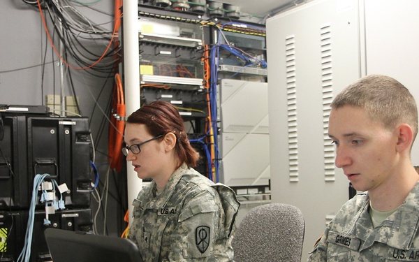 Two U.S. Army soldiers in ACU (Army Combat Uniform) camouflage are working at a desk with laptops in what appears to be a military communications or IT operations room. The soldier in the foreground has a name tape reading 'GRIMES' and both wear 'U.S. ARMY' chest tape along with matching unit shoulder patches. The background contains server racks, networking equipment, and extensive cable management infrastructure.