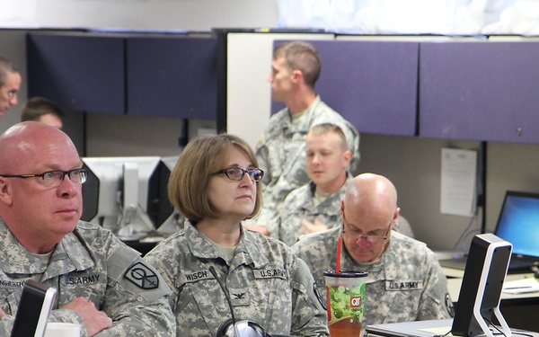 A group of U.S. Army personnel in ACU (Army Combat Uniform) camouflage are gathered in what appears to be a military operations or command center. Multiple soldiers are seated at workstations equipped with laptops and monitors, appearing to observe or attend a briefing. The environment resembles a tactical operations center (TOC) or training facility with cubicle-style dividers.