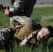 Two military personnel in MARPAT (Marine pattern) camouflage uniforms are engaged in what appears to be a combatives or martial arts training exercise on a grassy field. One service member is on the ground while the other applies a control technique, consistent with military close-quarters combat training. Both individuals are wearing standard-issue tactical gear including a plate carrier vest.