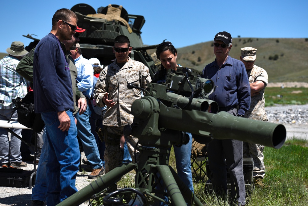 A U.S. Marine in desert MARPAT camouflage uniform is briefing a group of civilians on a crew-served weapon system, likely a TOW missile launcher or recoilless rifle mounted on a tripod. The setting appears to be an outdoor military demonstration or open house event on a range or training area. A military vehicle is visible in the background along with rolling hills under clear blue skies.