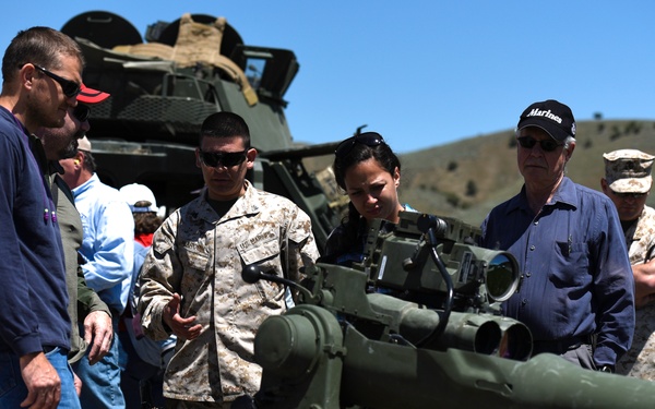A U.S. Marine in desert MARPAT camouflage uniform is briefing a group of civilians on a crew-served weapon system, likely a TOW missile launcher or recoilless rifle mounted on a tripod. The setting appears to be an outdoor military demonstration or open house event on a range or training area. A military vehicle is visible in the background along with rolling hills under clear blue skies.