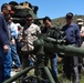A U.S. Marine in desert MARPAT camouflage uniform is briefing a group of civilians on a crew-served weapon system, likely a TOW missile launcher or recoilless rifle mounted on a tripod. The setting appears to be an outdoor military demonstration or open house event on a range or training area. A military vehicle is visible in the background along with rolling hills under clear blue skies.