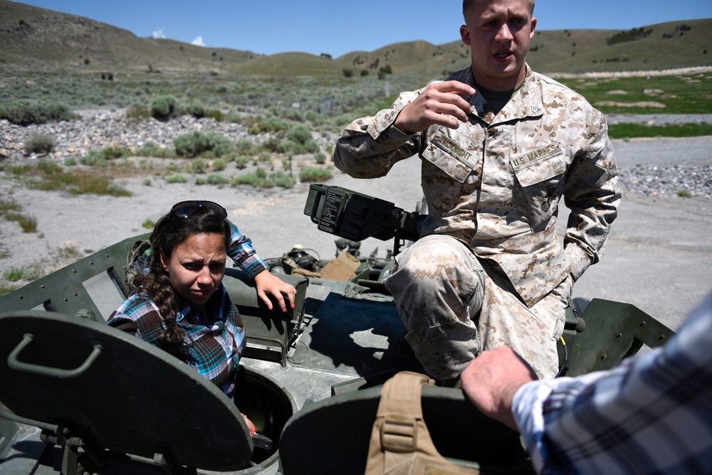 A U.S. Marines soldier in MARPAT desert camouflage uniform is explaining or demonstrating something on a military vehicle to a civilian woman in a plaid shirt. The setting appears to be an outdoor arid landscape with scrubland and hills in the background, suggesting a training area or public demonstration event. A third person's arm is partially visible in the foreground.