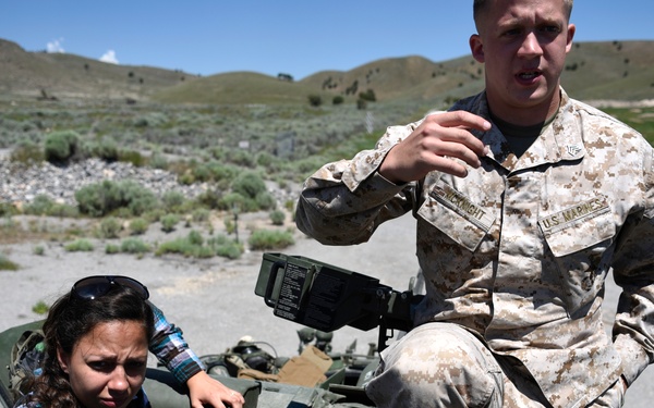 A U.S. Marines soldier in MARPAT desert camouflage uniform is explaining or demonstrating something on a military vehicle to a civilian woman in a plaid shirt. The setting appears to be an outdoor arid landscape with scrubland and hills in the background, suggesting a training area or public demonstration event. A third person's arm is partially visible in the foreground.
