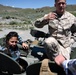 A U.S. Marines soldier in MARPAT desert camouflage uniform is explaining or demonstrating something on a military vehicle to a civilian woman in a plaid shirt. The setting appears to be an outdoor arid landscape with scrubland and hills in the background, suggesting a training area or public demonstration event. A third person's arm is partially visible in the foreground.