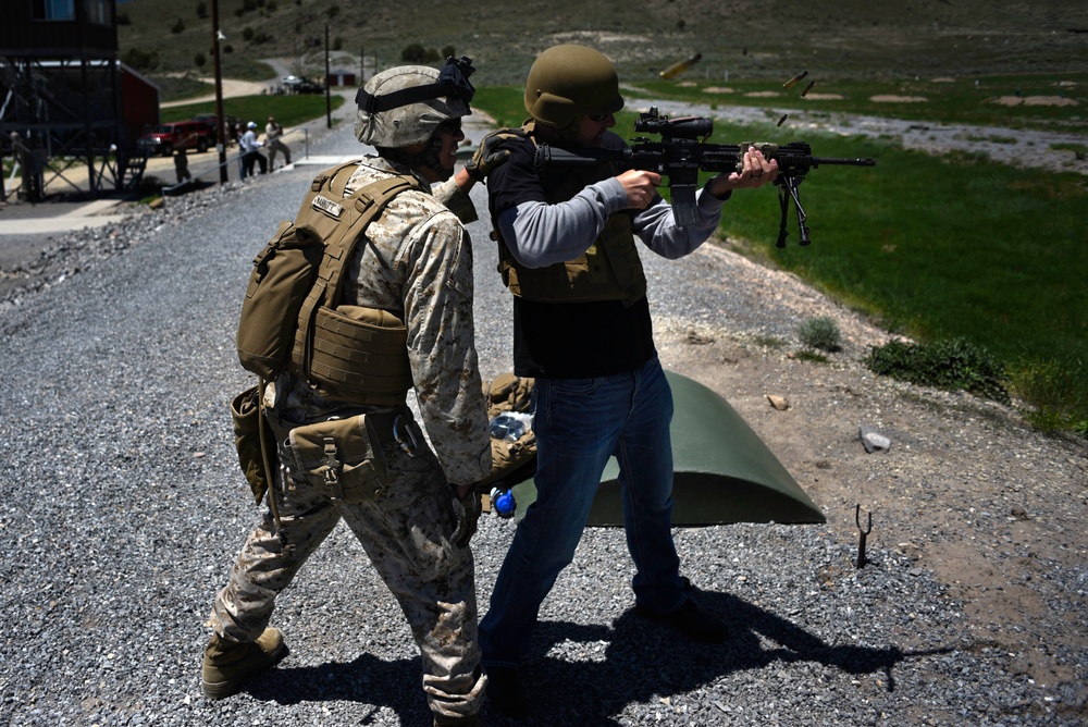 A military training exercise is taking place at an outdoor shooting range. A Marine in full combat uniform and gear is instructing or supervising a civilian wearing a ballistic vest and helmet who is firing a rifle with a bipod attachment. A spent casing is visibly ejecting from the weapon, indicating active firing.