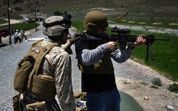 A military training exercise is taking place at an outdoor shooting range. A Marine in full combat uniform and gear is instructing or supervising a civilian wearing a ballistic vest and helmet who is firing a rifle with a bipod attachment. A spent casing is visibly ejecting from the weapon, indicating active firing.