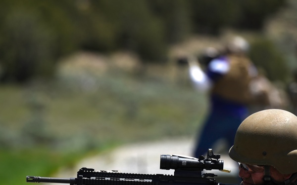A person wearing a ballistic helmet and tactical vest is in a prone/seated shooting position on a gravel surface, aiming a rifle equipped with an optic scope and bipod. Additional individuals are visible in the blurred background, suggesting a group training or range activity. The setting appears to be an outdoor shooting range or training area with natural vegetation in the background.