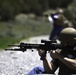 A person wearing a ballistic helmet and tactical vest is in a prone/seated shooting position on a gravel surface, aiming a rifle equipped with an optic scope and bipod. Additional individuals are visible in the blurred background, suggesting a group training or range activity. The setting appears to be an outdoor shooting range or training area with natural vegetation in the background.