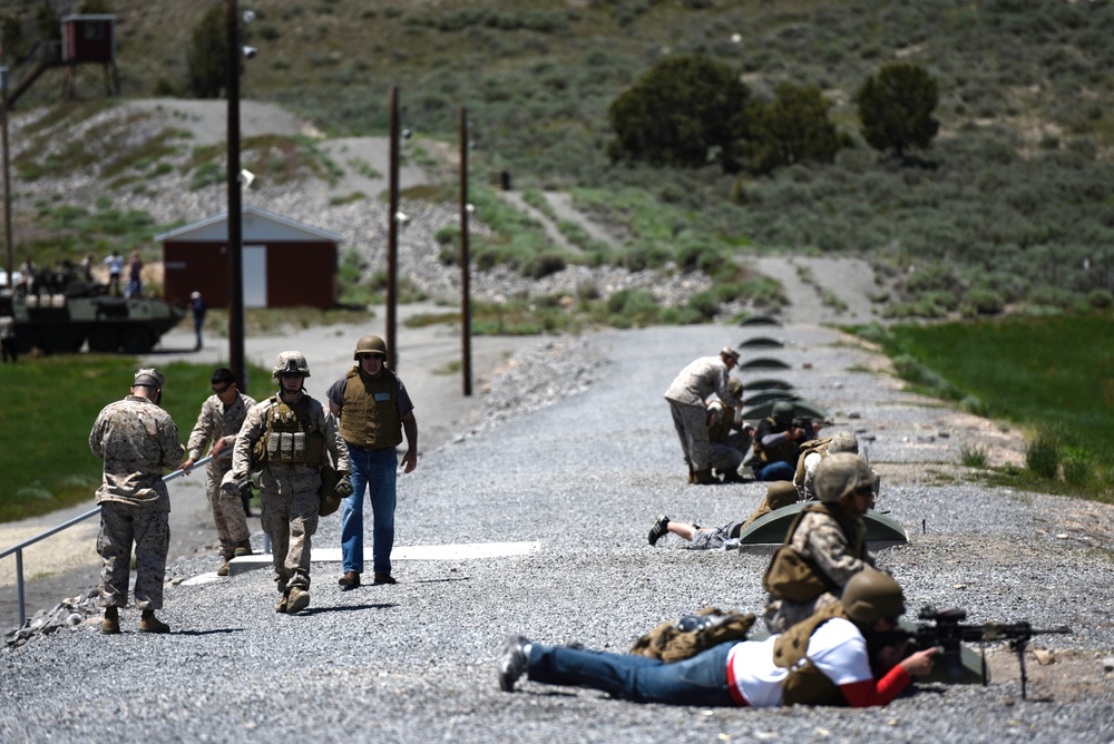 The image shows what appears to be a military training exercise or civilian firearms familiarization event at an outdoor range facility. Several individuals in military uniforms and civilian clothing are present, with some in prone shooting positions on a gravel road while others stand or walk nearby. Military vehicles are visible in the background along with range infrastructure.