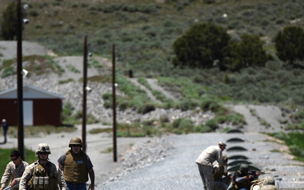 The image shows what appears to be a military training exercise or civilian firearms familiarization event at an outdoor range facility. Several individuals in military uniforms and civilian clothing are present, with some in prone shooting positions on a gravel road while others stand or walk nearby. Military vehicles are visible in the background along with range infrastructure.