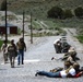 The image shows what appears to be a military training exercise or civilian firearms familiarization event at an outdoor range facility. Several individuals in military uniforms and civilian clothing are present, with some in prone shooting positions on a gravel road while others stand or walk nearby. Military vehicles are visible in the background along with range infrastructure.