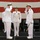 A formal military change of command ceremony is taking place on a stage in front of a large American flag backdrop. Three officers in white dress uniforms are prominently featured, with two exchanging salutes while a senior officer with admiral insignia stands at attention between them. A fourth uniformed personnel is visible in the background.