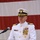 A senior United States Coast Guard officer in white dress uniform is speaking at a podium during a formal ceremony. The podium bears the official United States Coast Guard seal with the founding year 1790. An American flag is displayed prominently in the background.