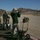 An outdoor rifle shooting range in a desert environment, likely in the American Southwest based on the arid landscape and mountain backdrop. Multiple shooters are positioned at numbered firing line stations, with the foreground shooter actively aiming a rifle downrange. A red range flag is visible in the distance, indicating the range is active.